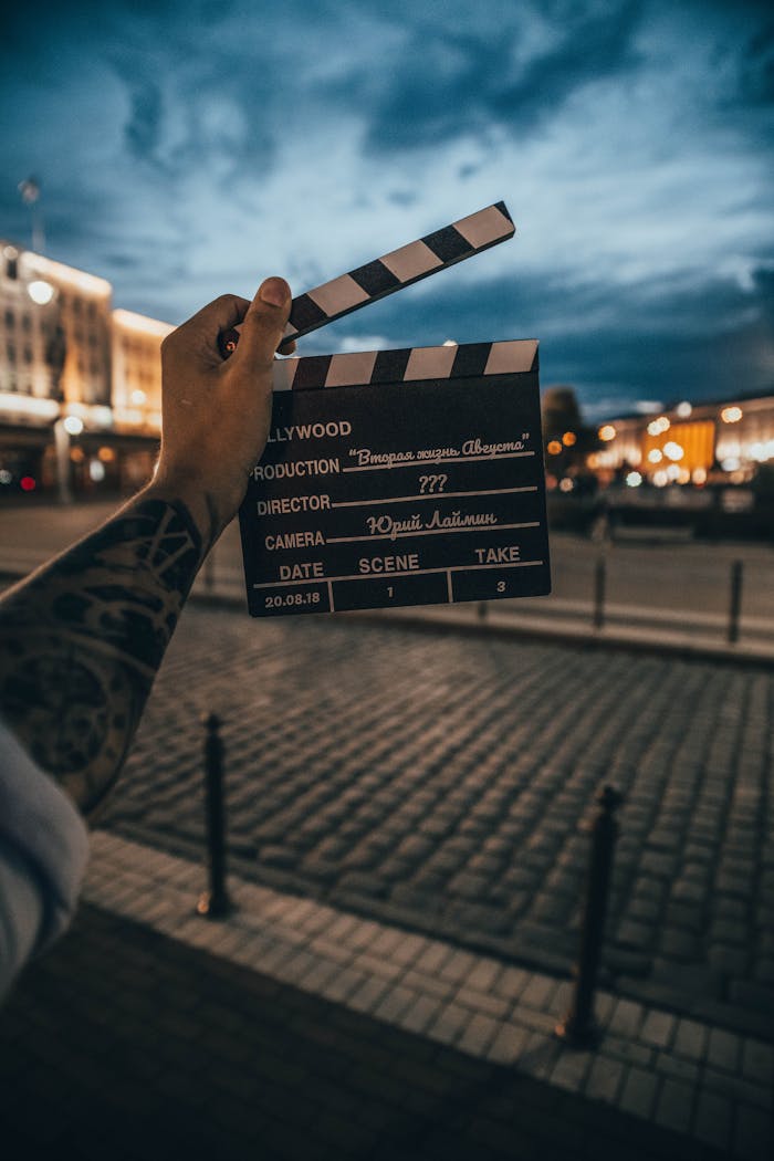 A tattooed hand holds a clapperboard during dusk on a city street, evoking cinematic vibes.